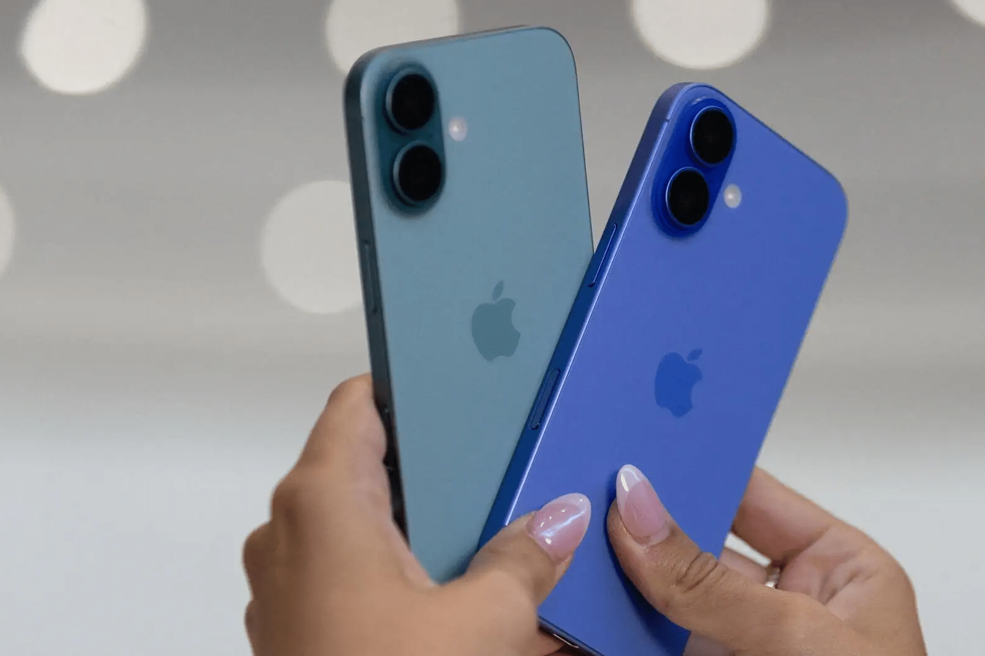 Two hands holding two different colored iPhones, one blue-green and one blue, both showing the Apple logo and dual rear cameras—devices like these are often targeted by stolen iPhone smuggling rings—against a blurred light background.