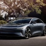A sleek, silver electric sedan is parked on a sunny street surrounded by greenery and modern buildings, reflecting sunlight off its smooth, aerodynamic body and black-trimmed wheels.