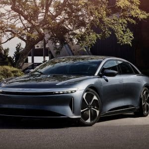 A sleek, silver electric sedan is parked on a sunny street surrounded by greenery and modern buildings, reflecting sunlight off its smooth, aerodynamic body and black-trimmed wheels.