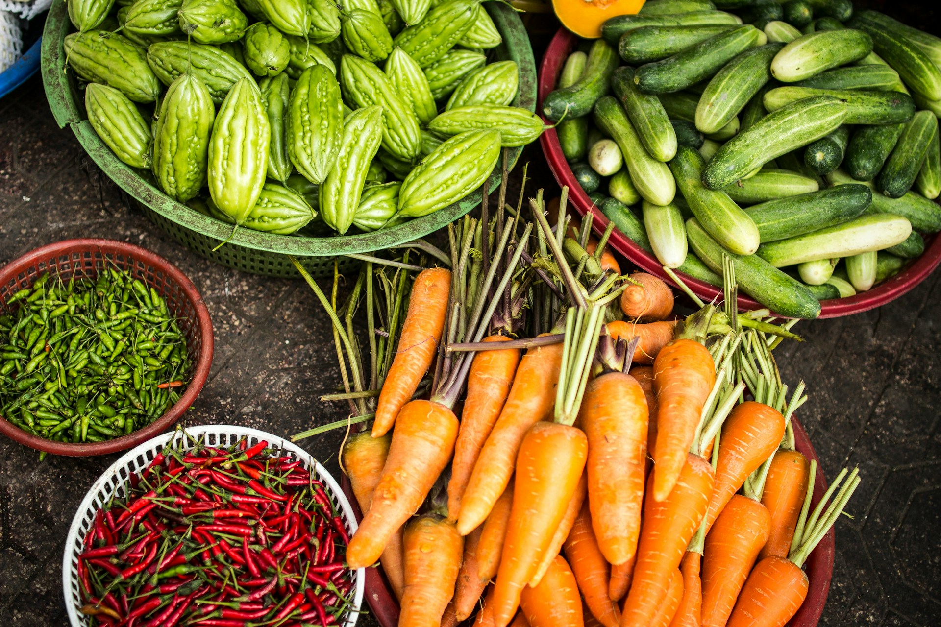 An assortment of fresh vegetables in baskets, including carrots, cucumbers, bitter melons, green chilies, and red chilies, displayed on a market floor highlights the importance of food plant sanitation services for maintaining freshness and safety.