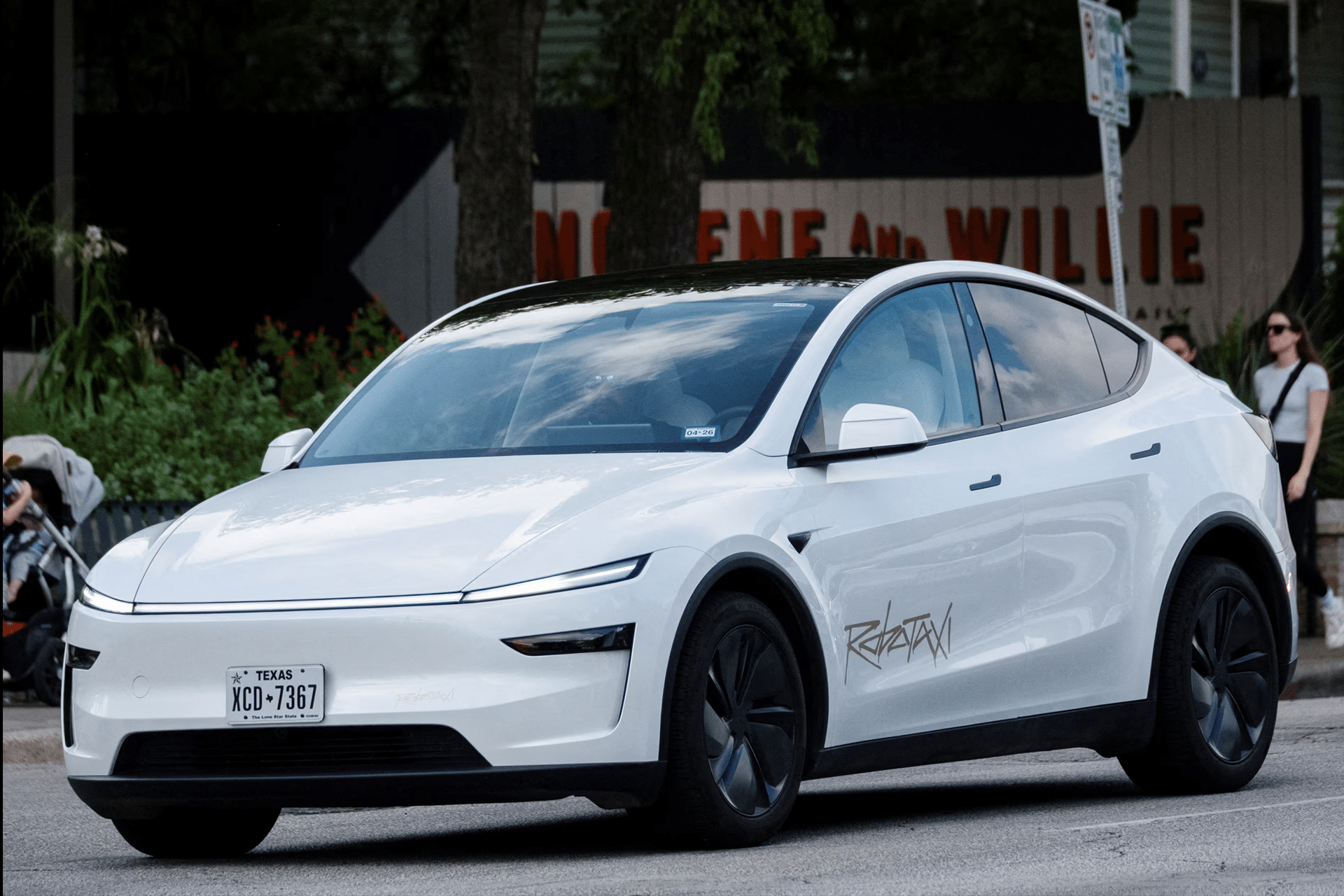 A sleek white electric car with tinted windows and black wheel covers is parked on a street. Featuring a modern design inspired by Elon Musk robotaxi production, it sports "Ratapan" on the side and a Texas license plate, with people and greenery nearby.