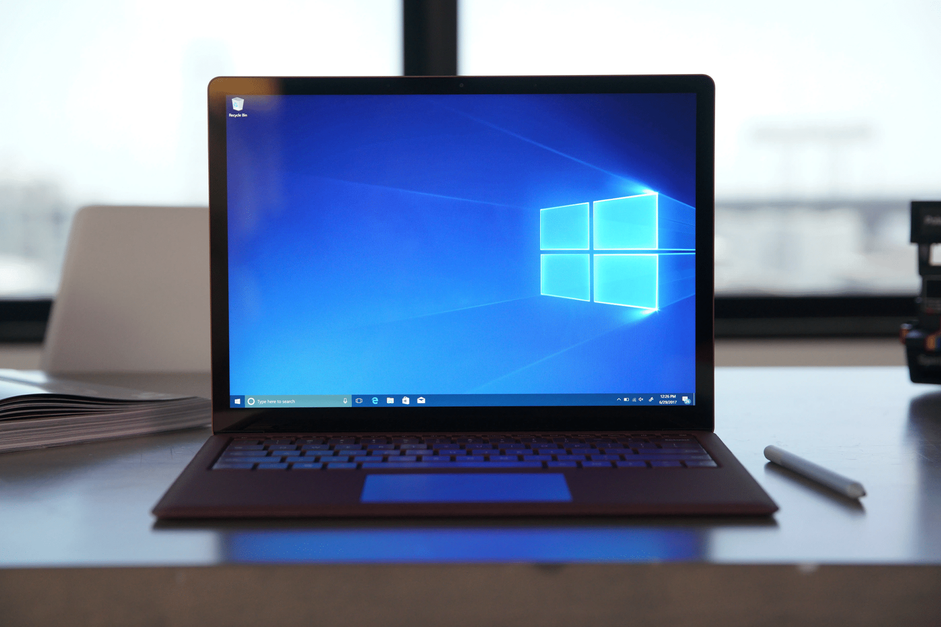 A laptop on a desk displays the Windows 10 home screen, hinting at an upcoming Windows 10 end of life and a possible switch to Mac. Nearby are an open notebook, a pen, and books, with natural light streaming in through the window.