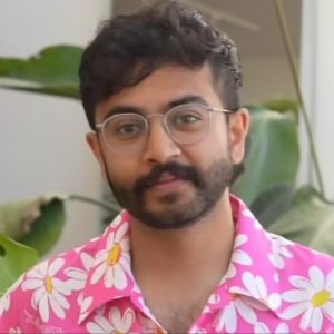 A person with glasses and a beard stands indoors, wearing a bright pink shirt with white and yellow daisy prints. Green plants are visible in the background, hinting at a fresh start—much like when Abidur Chowdhury leaves Apple.