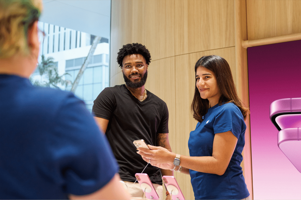 A man and a woman stand at a store counter, possibly discussing Apple sales restructuring with an employee. The man is holding a smartphone while the woman in a blue shirt smiles, also holding her phone. Another person is visible from behind.