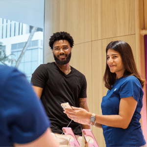 A man and a woman stand at a store counter, possibly discussing Apple sales restructuring with an employee. The man is holding a smartphone while the woman in a blue shirt smiles, also holding her phone. Another person is visible from behind.