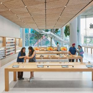A modern Apple Store interior at the Apple Noida store in DLF Mall of India features large windows, display tables with devices, customers browsing and staff assisting. Greenery and a staircase complete the bright, open space.