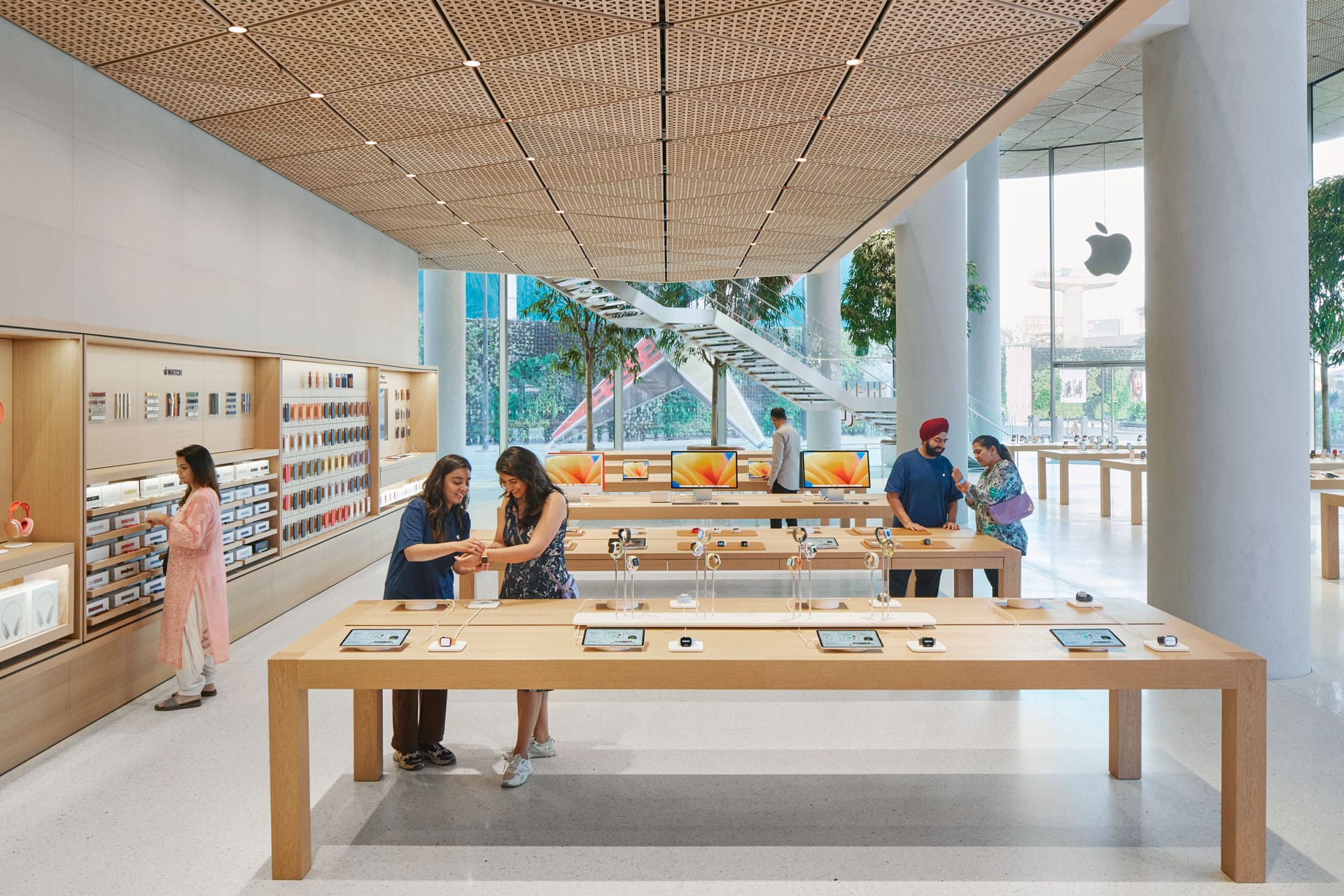 A modern Apple Store interior at the Apple Noida store in DLF Mall of India features large windows, display tables with devices, customers browsing and staff assisting. Greenery and a staircase complete the bright, open space.