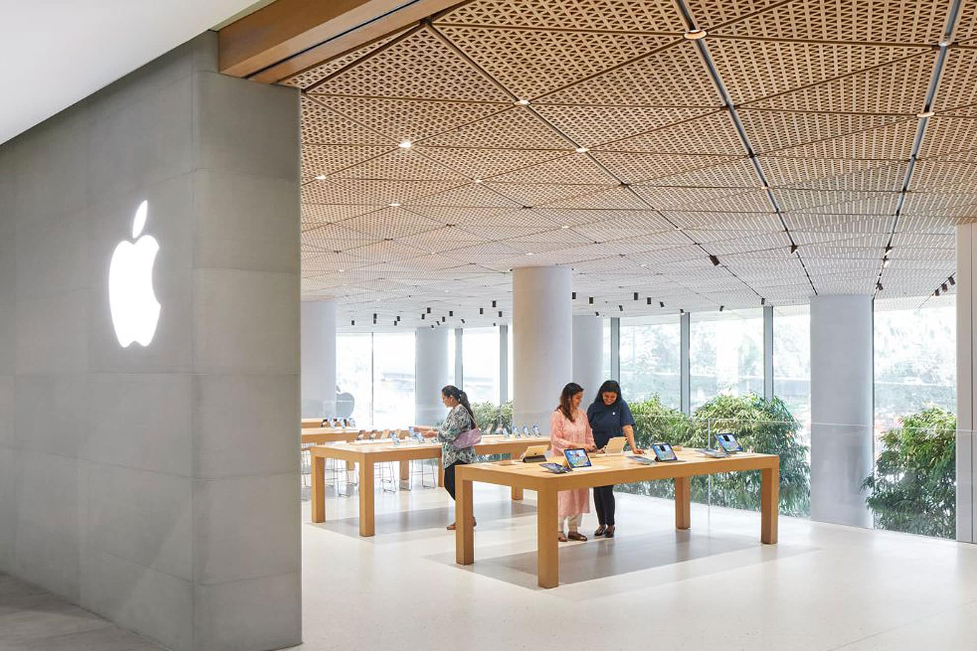 Three people stand at wooden tables displaying electronics inside the new Apple Noida store at DLF Mall of India, featuring a bright interior with large windows and a prominent white Apple logo on the wall during the Apple store opening.