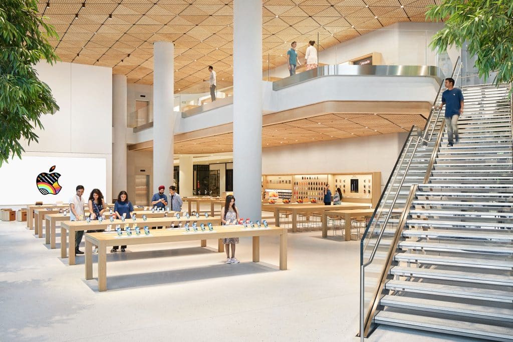 Spacious Apple Noida store at DLF Mall of India with high ceilings, wooden tables displaying devices, customers browsing, a large staircase, colorful Apple logo on a screen, and green plants inside the modern, bright interior.
