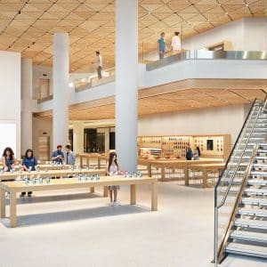 Spacious Apple Noida store at DLF Mall of India with high ceilings, wooden tables displaying devices, customers browsing, a large staircase, colorful Apple logo on a screen, and green plants inside the modern, bright interior.