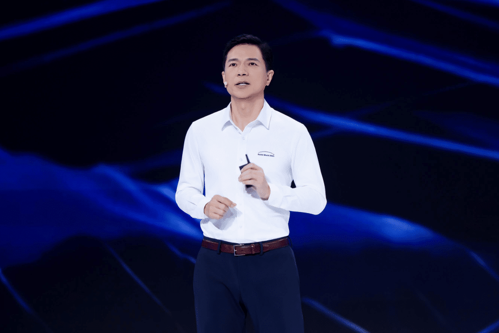 A man in a white dress shirt and dark pants stands on a stage with a blue abstract background, speaking and holding a small device in his right hand, possibly presenting the new Baidu M100 or M300 chips.