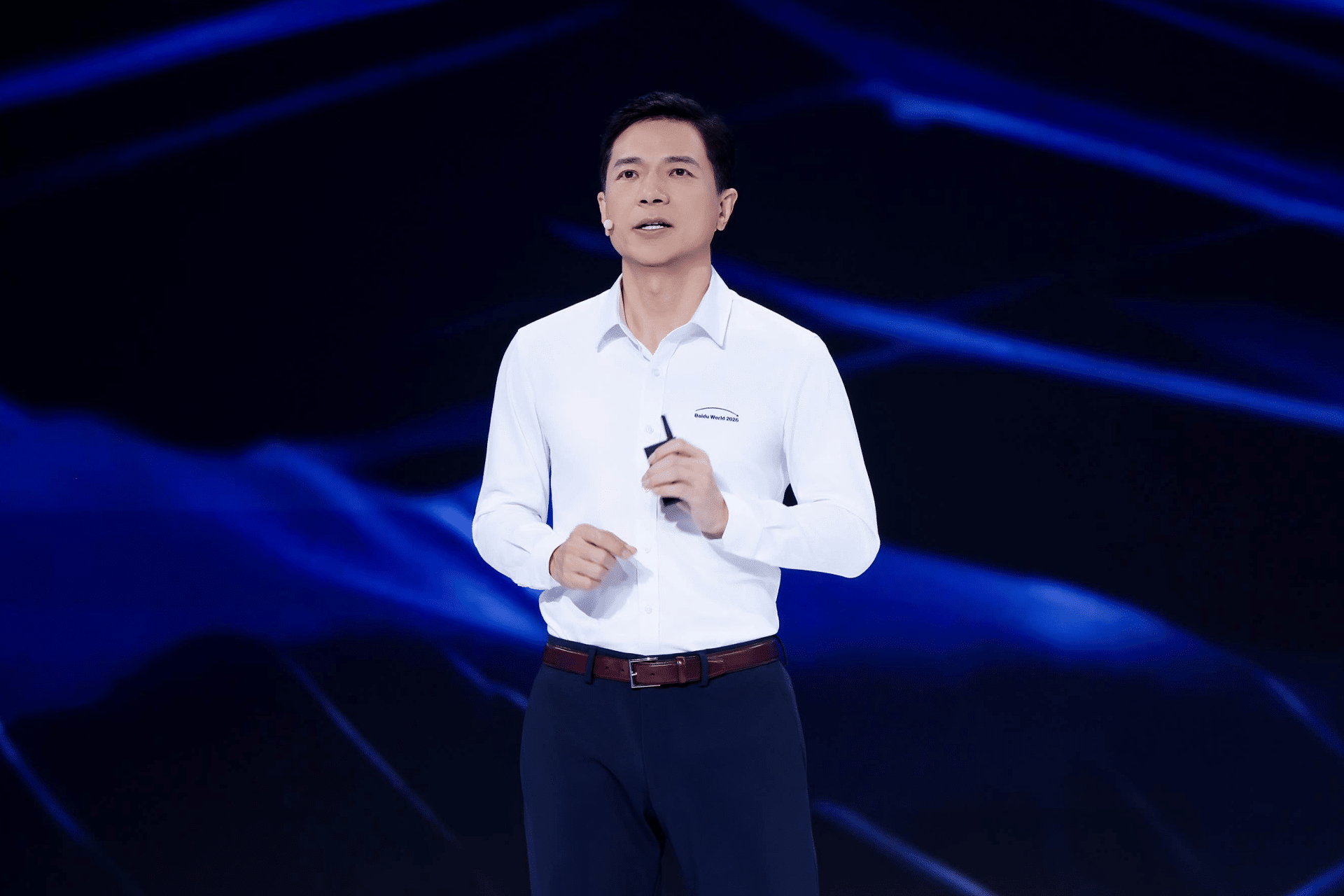 A man in a white dress shirt and dark pants stands on a stage with a blue abstract background, speaking and holding a small device in his right hand, possibly presenting the new Baidu M100 or M300 chips.