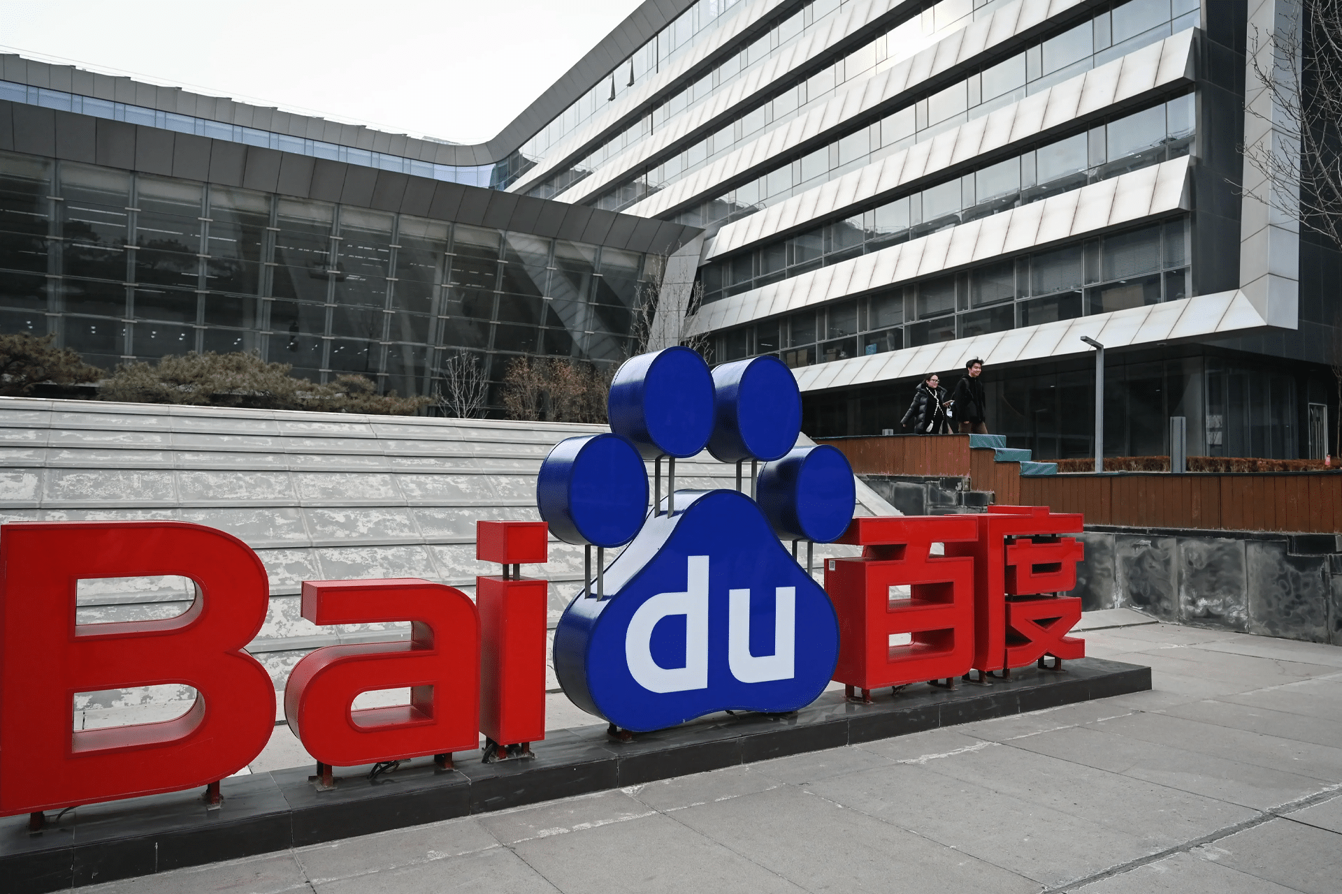 Large Baidu logo with red, blue, and white colors stands in front of a modern glass office building in China. A person walks on a pathway above the sign, highlighting Baidu's role in AI chips innovation.