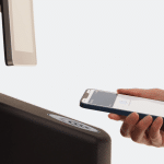 A person holds a smartphone near a payment terminal, using Apple Wallet to make a contactless transaction with their digital passport. The background is white and minimalistic.