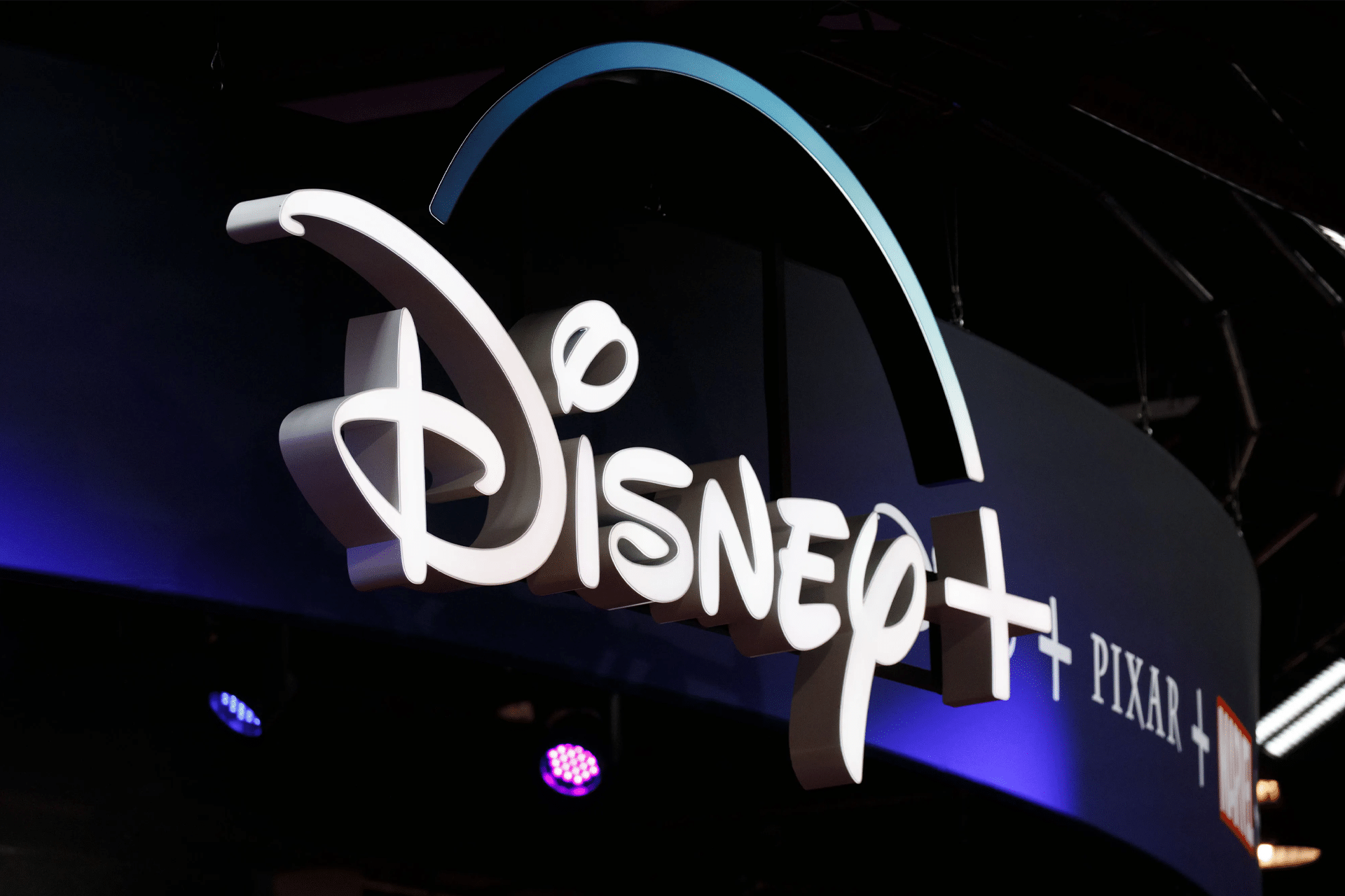 The Disney+ logo is illuminated on a dark background, with the Pixar and Marvel logos partially visible to the right. Purple and blue lights shine beneath the display, highlighting Disney Plus on older Apple devices.