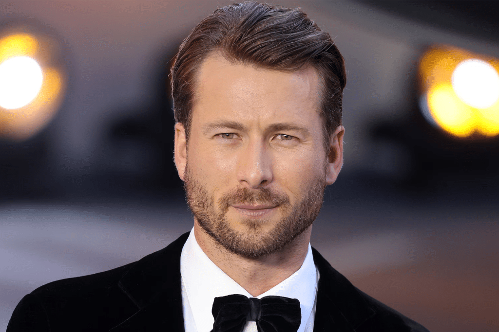 A man with short, slicked-back brown hair and a trimmed beard wears a black tuxedo with a bow tie, standing before bright, out-of-focus lights at the Apple Original Films The Flick premiere.