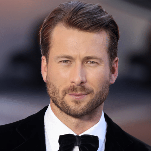 A man with short, slicked-back brown hair and a trimmed beard wears a black tuxedo with a bow tie, standing before bright, out-of-focus lights at the Apple Original Films The Flick premiere.