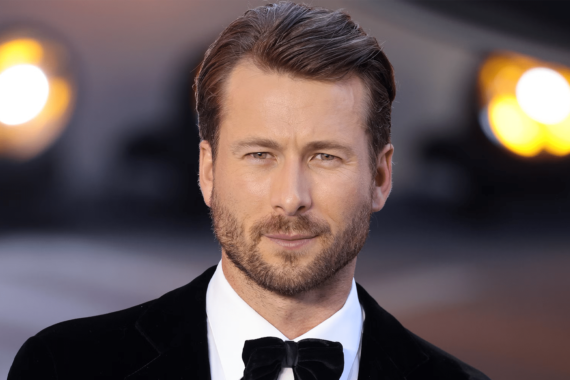 A man with short, slicked-back brown hair and a trimmed beard wears a black tuxedo with a bow tie, standing before bright, out-of-focus lights at the Apple Original Films The Flick premiere.