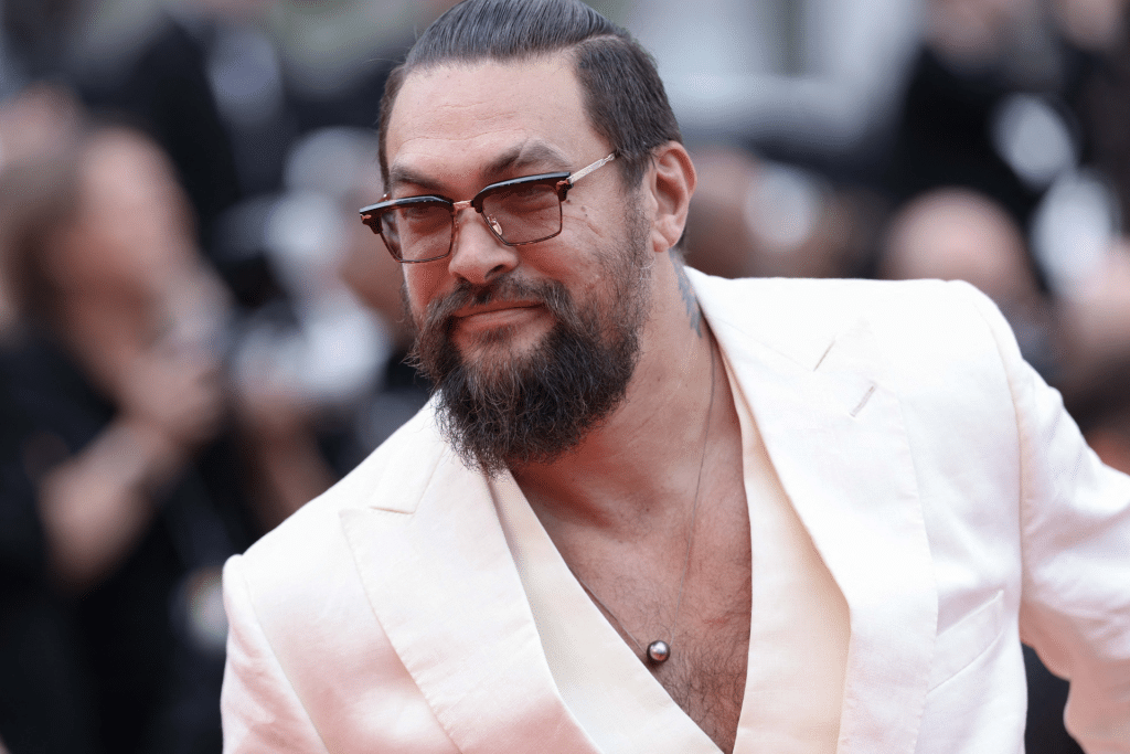 A man with long dark hair tied back, wearing sunglasses and a white suit with an open collar, smiles while posing at a formal event—reminiscent of Jason Momoa's charismatic style in his popular biker series—with blurred people and cameras in the background.