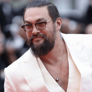 A man with long dark hair tied back, wearing sunglasses and a white suit with an open collar, smiles while posing at a formal event—reminiscent of Jason Momoa's charismatic style in his popular biker series—with blurred people and cameras in the background.