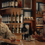 Two men sit at a bar counter having a conversation, with wine bottles displayed on shelves behind them. One man wears glasses and a light jacket, while the other gestures while discussing an OpenAI hardware device. Drinks and cups are on the counter.