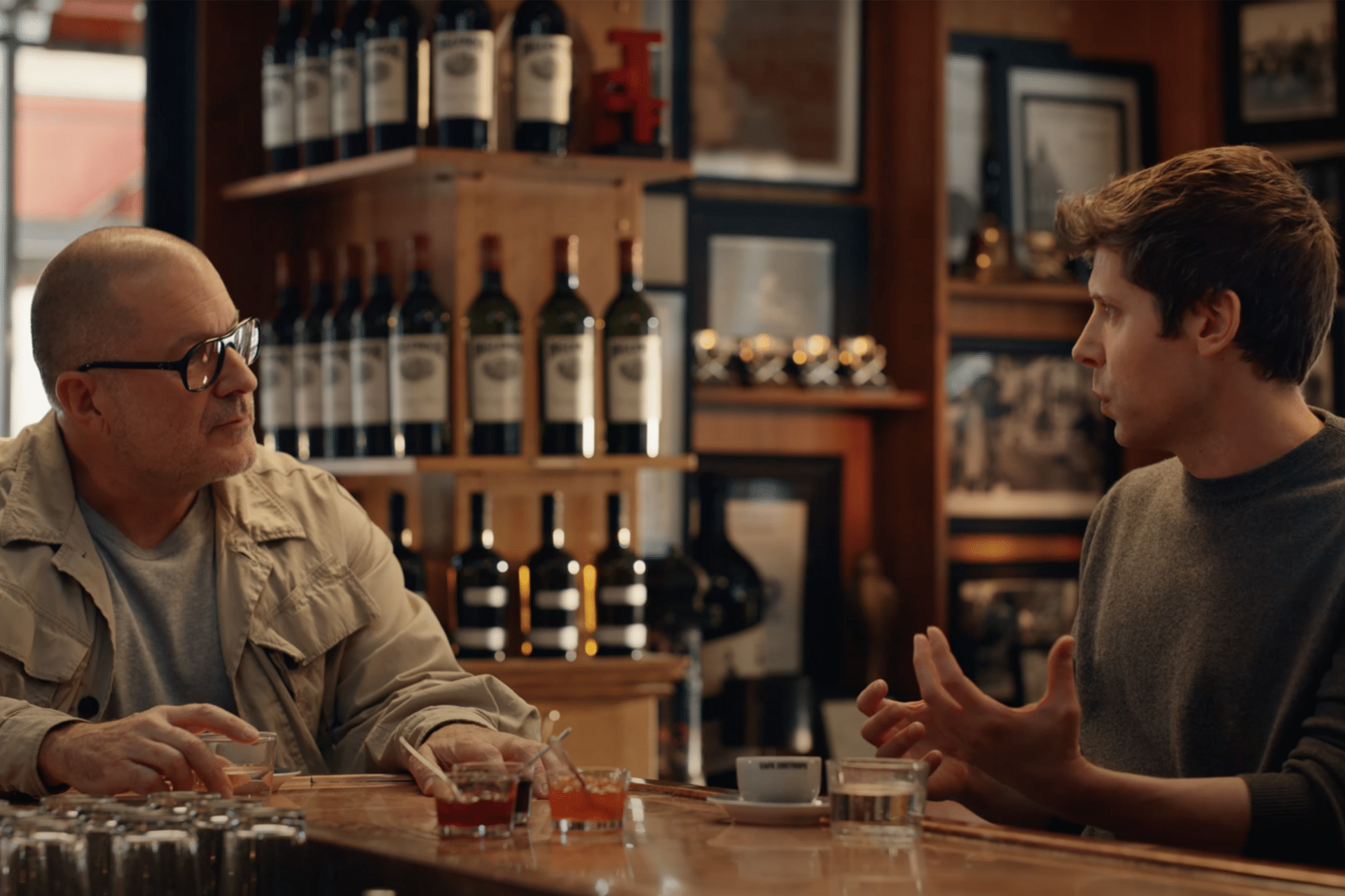 Two men sit at a bar counter having a conversation, with wine bottles displayed on shelves behind them. One man wears glasses and a light jacket, while the other gestures while discussing an OpenAI hardware device. Drinks and cups are on the counter.