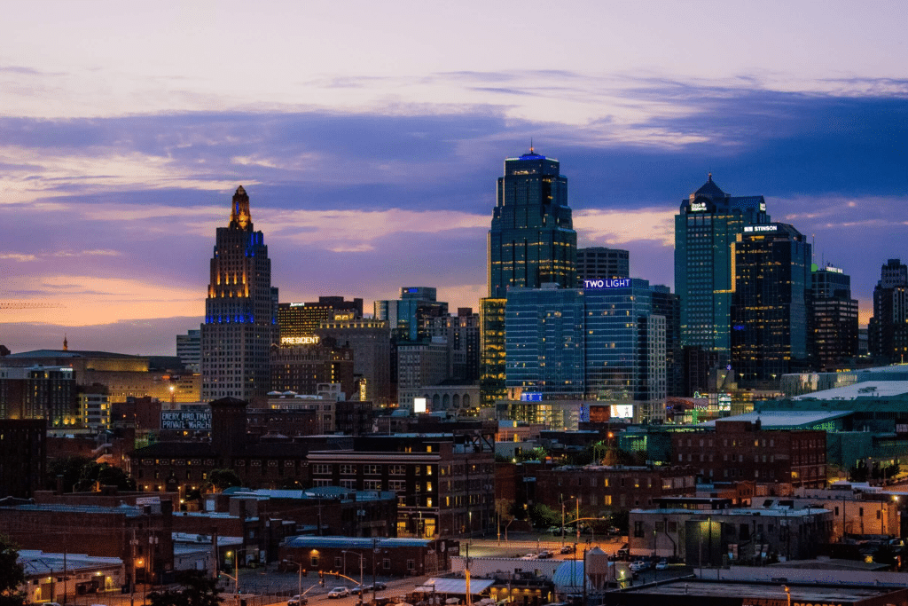 City skyline at dusk with tall buildings lit up, including a historic tower and modern glass skyscrapers, against a colorful sunset sky with purple and orange hues—captured perfectly in Apple Maps Kansas City. Industrial and residential buildings are visible in the foreground.