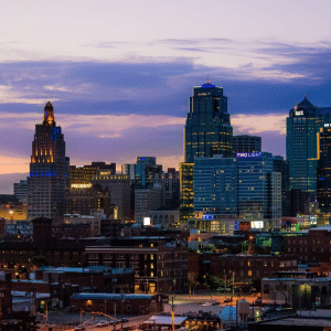 City skyline at dusk with tall buildings lit up, including a historic tower and modern glass skyscrapers, against a colorful sunset sky with purple and orange hues—captured perfectly in Apple Maps Kansas City. Industrial and residential buildings are visible in the foreground.