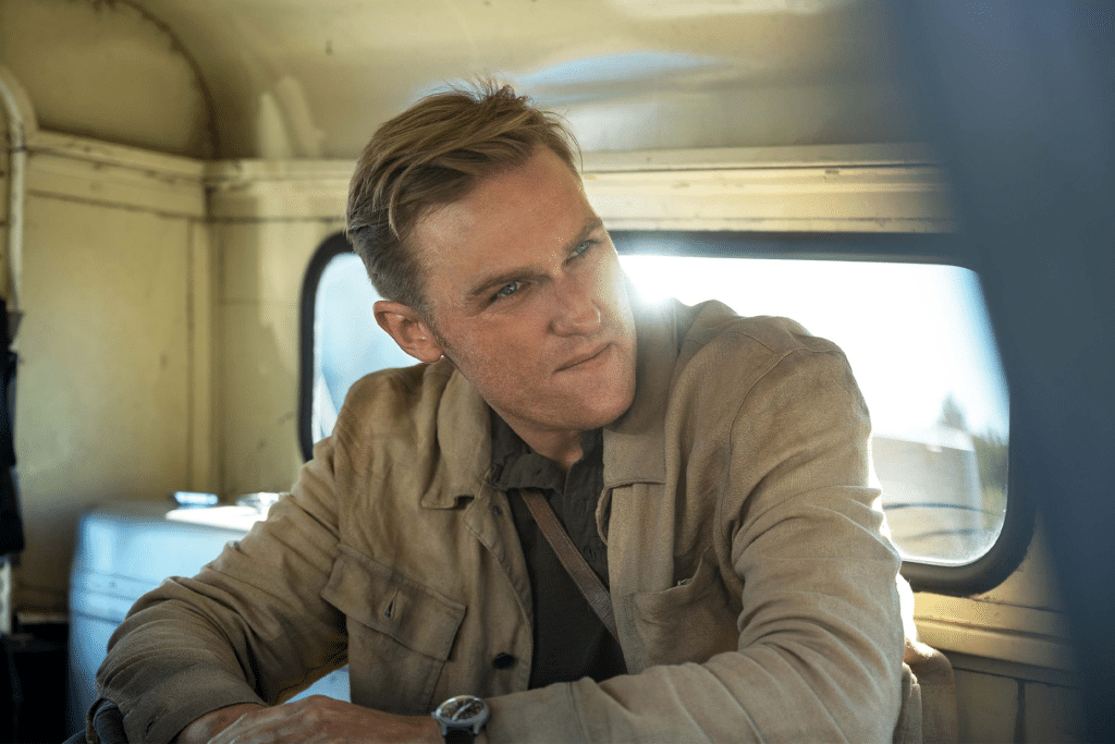 Wyatt Russell, with short blond hair, wearing a light brown jacket and dark shirt, sits inside an old vehicle from Monarch, looking to his left as sunlight streams through the window behind him.