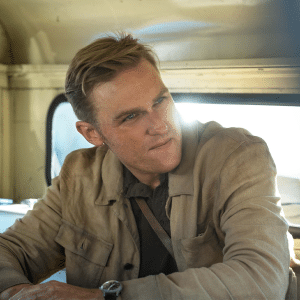 Wyatt Russell, with short blond hair, wearing a light brown jacket and dark shirt, sits inside an old vehicle from Monarch, looking to his left as sunlight streams through the window behind him.