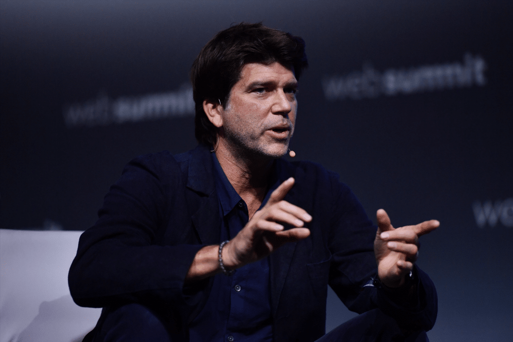 A man with dark hair sits and gestures with his hands while speaking on stage at Web Summit 2025, wearing a dark jacket and shirt, with a headset microphone. The blurred background references Apple Music.