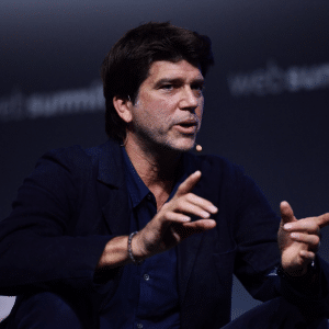 A man with dark hair sits and gestures with his hands while speaking on stage at Web Summit 2025, wearing a dark jacket and shirt, with a headset microphone. The blurred background references Apple Music.