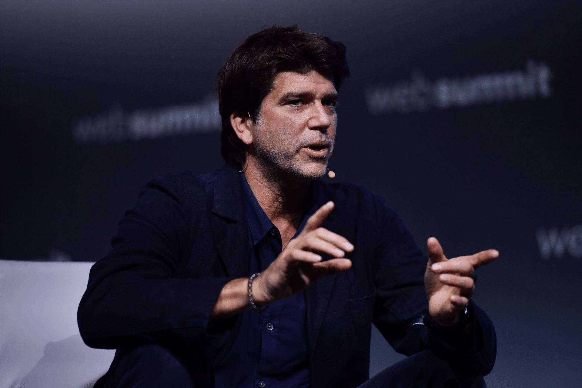 A man with dark hair sits and gestures with his hands while speaking on stage at Web Summit 2025, wearing a dark jacket and shirt, with a headset microphone. The blurred background references Apple Music.
