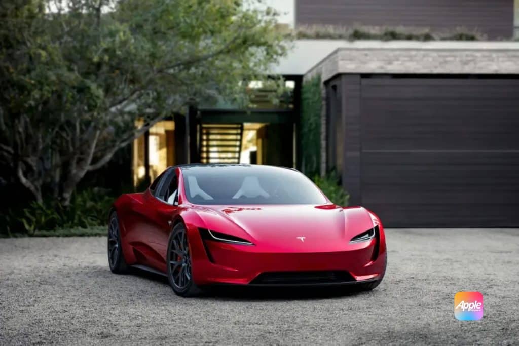 A sleek red Tesla sports car, inspired by Elon Musk’s vision, is parked on a gravel driveway in front of a modern house. The car's glossy finish reflects its surroundings. An Apple logo is visible in the bottom right corner.