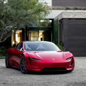 A sleek red Tesla sports car, inspired by Elon Musk’s vision, is parked on a gravel driveway in front of a modern house. The car's glossy finish reflects its surroundings. An Apple logo is visible in the bottom right corner.