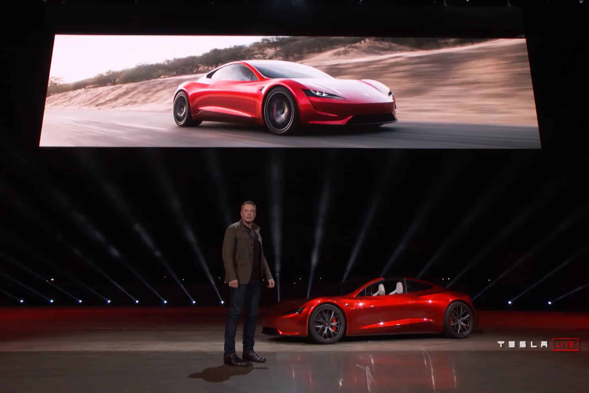 A man stands on stage beside a red Tesla sports car, with a large screen above showing the same car driving on a road. The stage is dimly lit with spotlights, and the Tesla logo appears in the corner. Elon Musk is presenting the scene.