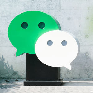 Two large WeChat logo sculptures, one green and one white, stand on a black pedestal outdoors against a concrete wall, with some green plants to the left—perhaps symbolizing the impact of the Apple WeChat deal.