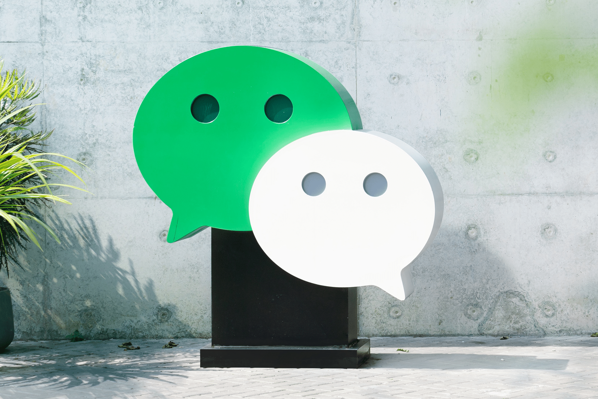 Two large WeChat logo sculptures, one green and one white, stand on a black pedestal outdoors against a concrete wall, with some green plants to the left—perhaps symbolizing the impact of the Apple WeChat deal.