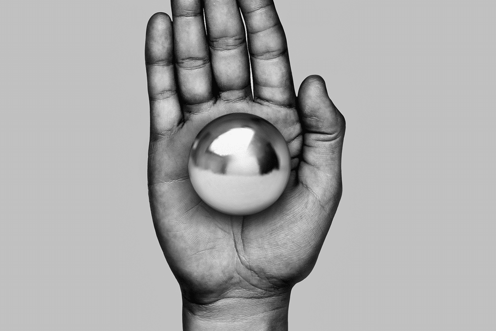 A grayscale photo of a hand held upright with the palm facing forward, holding a shiny metallic sphere—symbolizing innovation like X-Energy funding SMRs—centered in the palm against a plain gray background.