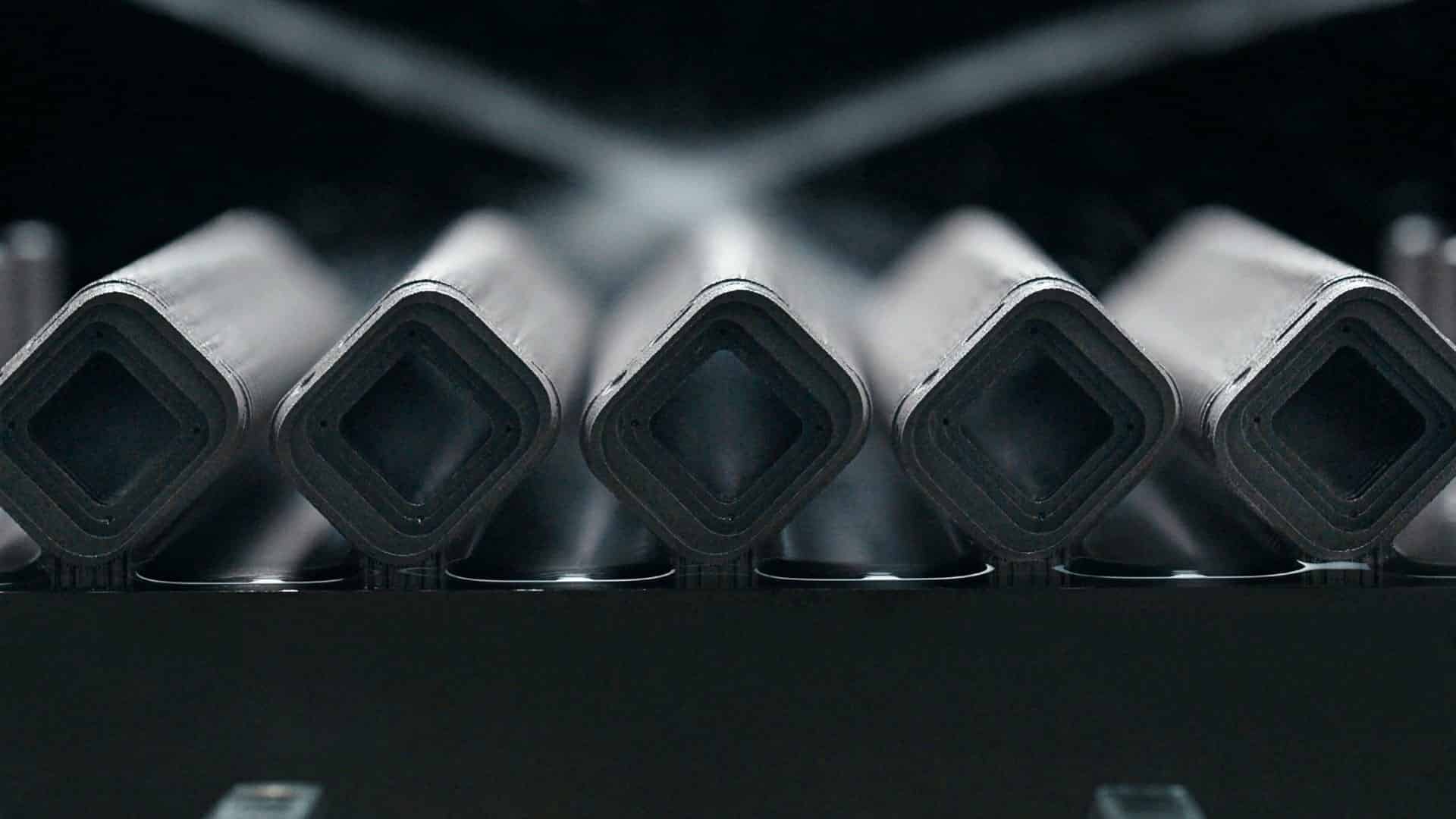 Close-up view of five hollow, square-shaped metal tubes arranged in a row, with water spray misting over them—evoking the precision found in 3D printing processes—in an industrial setting.