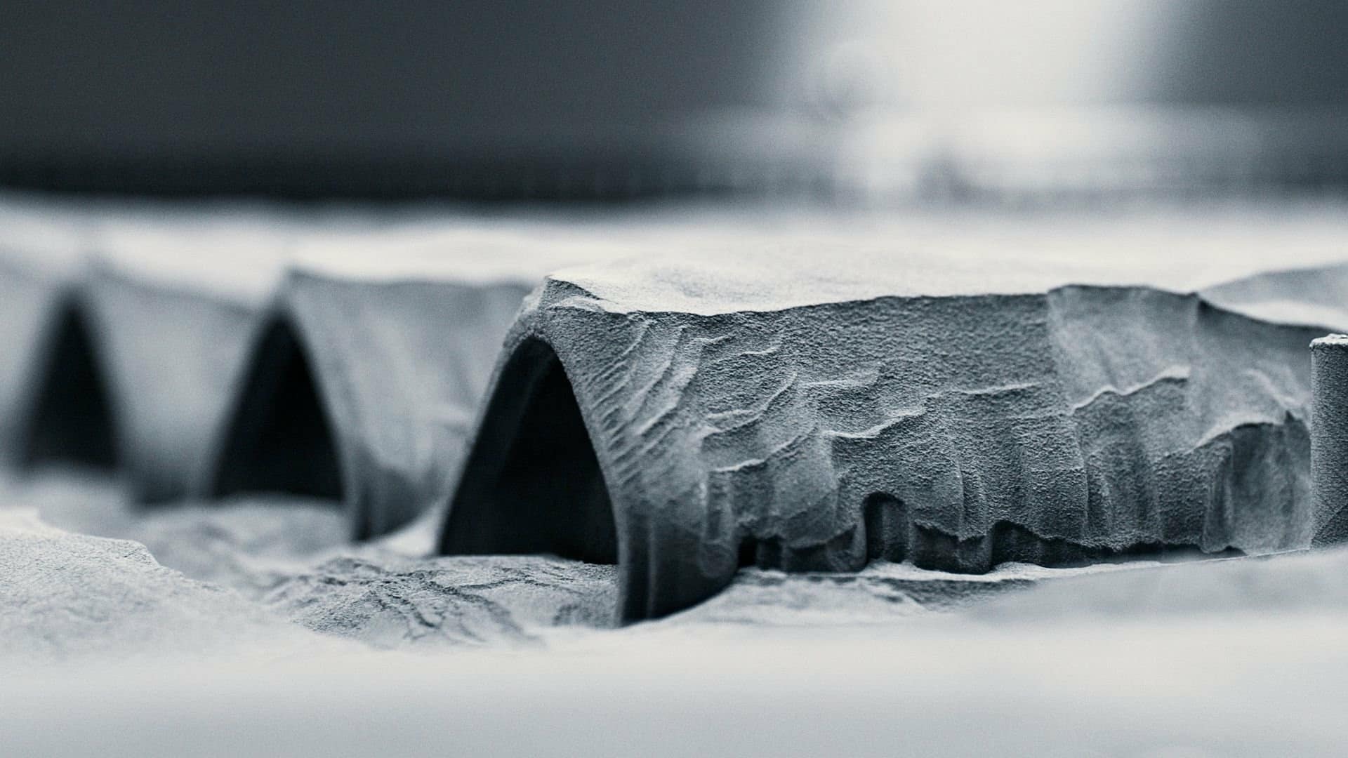Close-up of a detailed, miniature landscape resembling arched rock formations or cave entrances, with textured surfaces and fine dust or sand—crafted using 3D printing techniques—in grayscale tones.