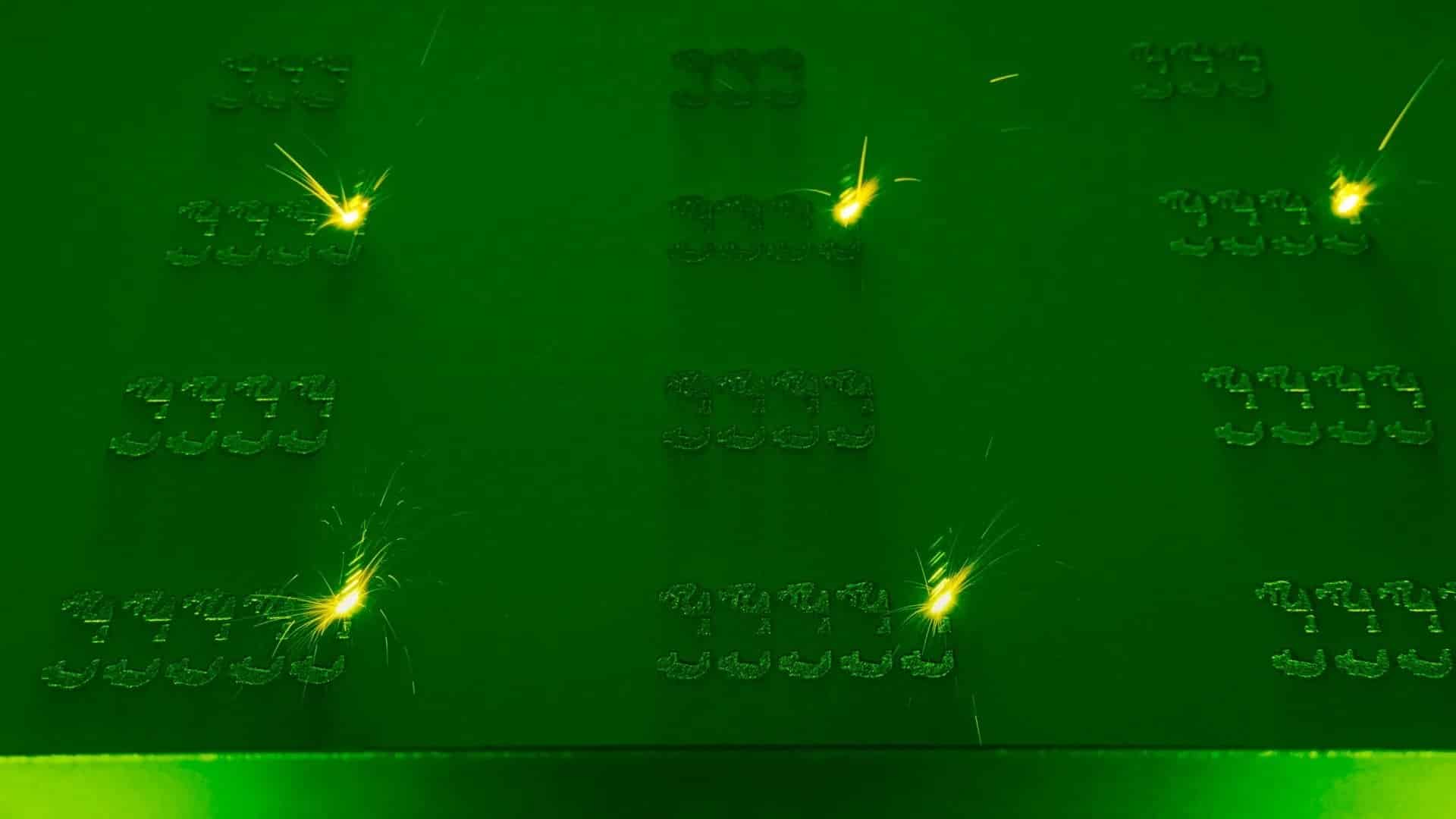 Bright yellow sparks appear at several points on a flat green metal surface, showing a 3D printing process in action as it creates small, repetitive geometric shapes in rows.