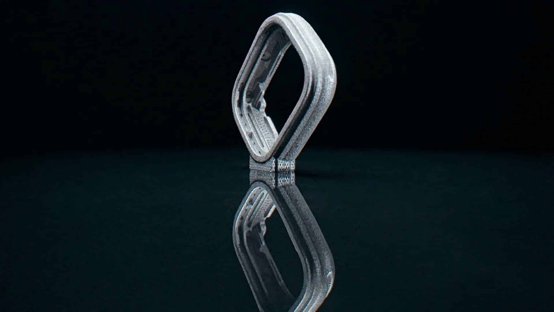 A metallic, octagonal smartwatch case—created with Apple 3D printing—stands upright on a glossy black surface, casting a clear reflection beneath it against a dark background.