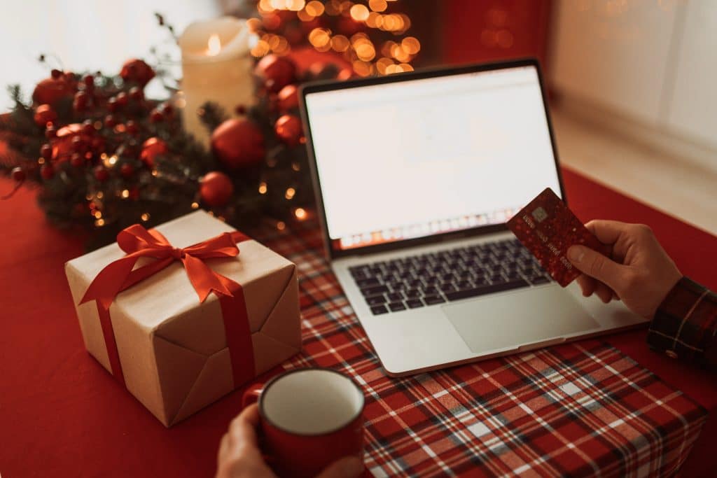 A person holding a credit card and a mug sits at a table with a wrapped gift, an open laptop, festive decorations, and warm lights in the background, ready for some seamless AI holiday shopping.