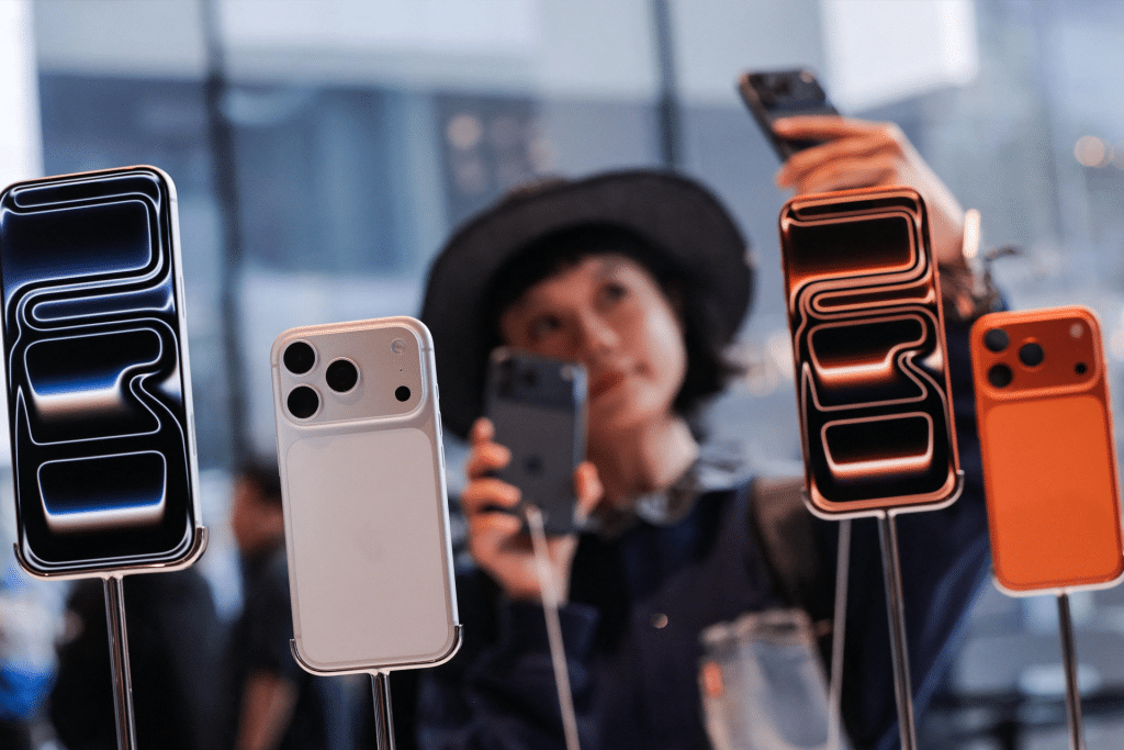 A person wearing a hat takes a selfie with an iPhone, while several colorful smartphones are displayed on stands in the foreground at a Singles Day tech event in China.