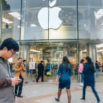People walk in front of a large Apple Store with a prominent Apple logo. A man in the foreground looks at his phone, possibly exploring third-party app stores on iOS 26.2. The store’s glass facade reveals more people and displays inside.