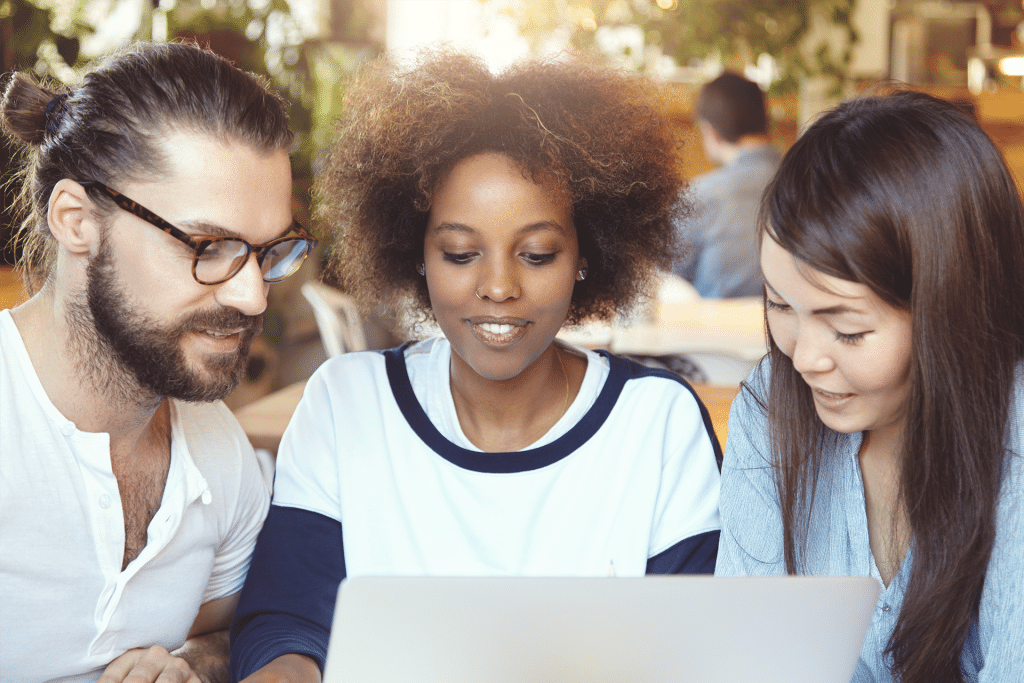 Three young adults sit closely together, smiling as they engage with AI in higher education on a laptop, appearing deeply involved in a collaborative activity within a bright, casual indoor setting.
