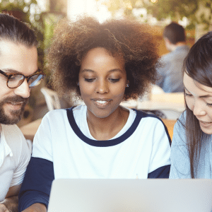 Three young adults sit closely together, smiling as they engage with AI in higher education on a laptop, appearing deeply involved in a collaborative activity within a bright, casual indoor setting.