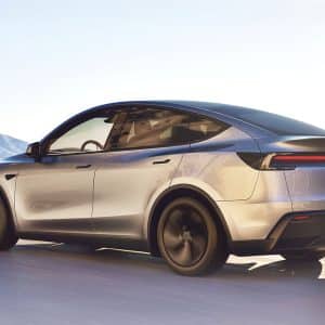 A sleek silver electric car with tinted windows and Musk-inspired aerodynamic design drives on a sunlit road, mountains and clear skies behind it—its thin horizontal taillights hinting at data-driven innovation.