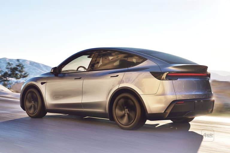A sleek silver electric car with tinted windows and Musk-inspired aerodynamic design drives on a sunlit road, mountains and clear skies behind it—its thin horizontal taillights hinting at data-driven innovation.